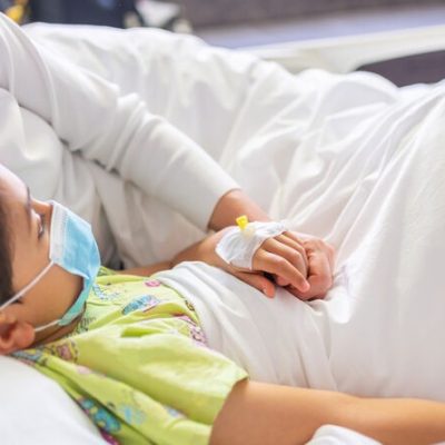 Young mother with protective face mask holding son's hand while lying on the hospital bed with him. Emotional Family Moment. Child vascular access